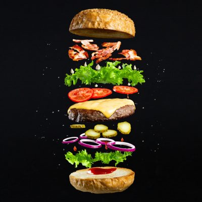 Floating burger isolated on black wooden background. Ingredients of a delicious burger with ground beef patty, lettuce, bacon, onions, tomatoes and cucumbers